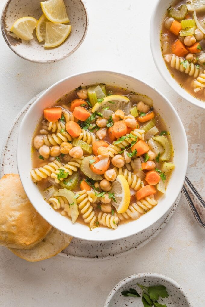 Overhead view of a table with several bowls of chickpea noodle soup, with garnishes of lemon and parsley, and biscuits.