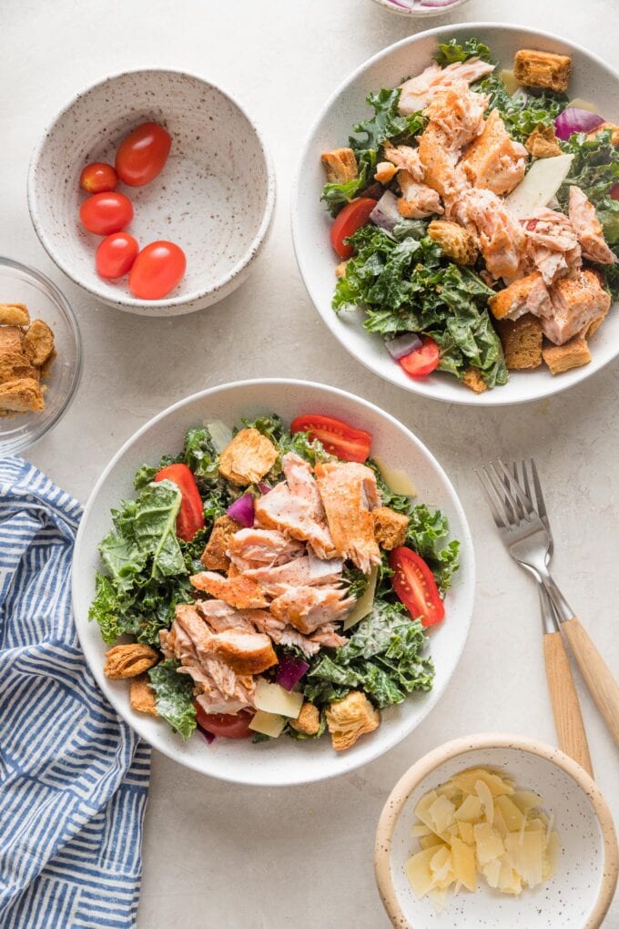 Two salad bowls filled with hearty lunch portions of salmon kale Caesar salads, with extra tomatoes and Parmesan in bowls on the side.