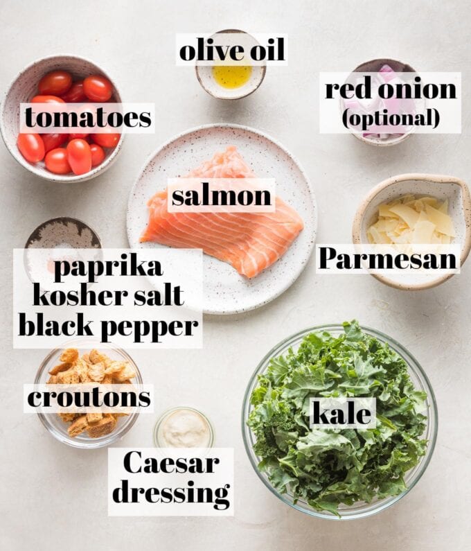 Labeled overhead photo of a salmon filet, olive oil, paprika, salt, and pepper, ready to cook, and prep bowls filled with curly kale, cherry tomatoes, shaved Parmesan, red onion, croutons, and Caesar salad dressing.