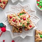 White star plate piled high with Christmas blondies studded with red and green M&Ms and chocolate chips, with extra M&Ms and Christmas decorations scattered on the counter in the background.