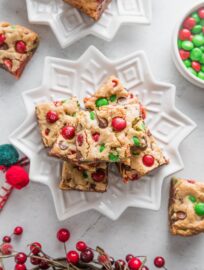White star plate piled high with Christmas blondies studded with red and green M&Ms and chocolate chips, with extra M&Ms and Christmas decorations scattered on the counter in the background.
