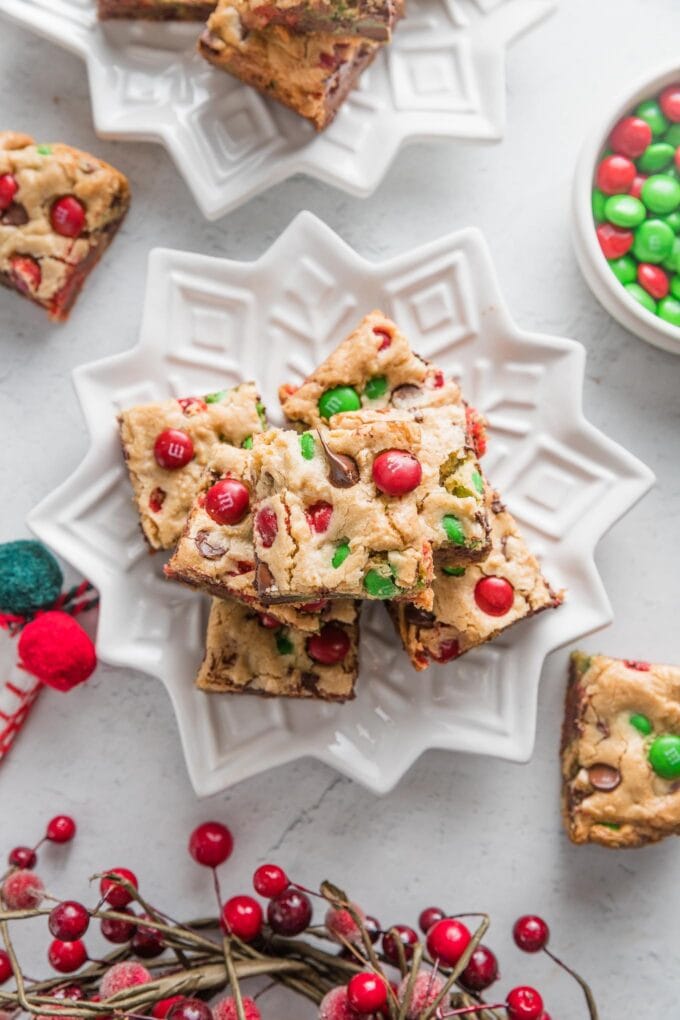 White star plate piled high with Christmas blondies studded with red and green M&Ms and chocolate chips, with extra M&Ms and Christmas decorations scattered on the counter in the background.