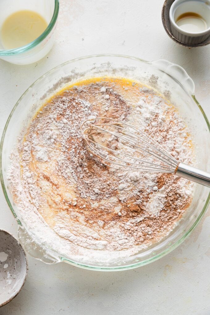 A whisk beginning to combine French toast batter in a shallow pie plate, with streaks of sugar and spices still visible.