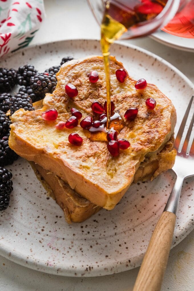 Close up of maple syrup being poured over eggnog French toast with pomegranate arils on top.