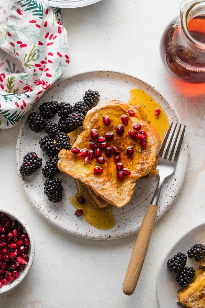 Overhead view of a small white ceramic plate with two slices of eggnog French toast served with maple syrup, blackberries, and pomegranate seeds.