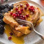 Close up of a fork pulling off bites of eggnog French toast from a stack of slices on a small plate.