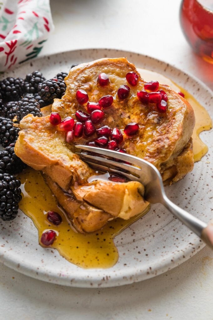 Close up of a fork pulling off bites of eggnog French toast from a stack of slices on a small plate.