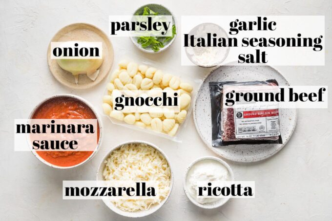 Labeled overhead photo showing a package of ground beef, shelf stable potato gnocchi, marinara sauce, shredded mozzarella, ricotta cheese, half of a yellow onion, fresh parsley, minced garlic, Italian seasoning, and salt all measured into plates and prep bowls and ready to cook.
