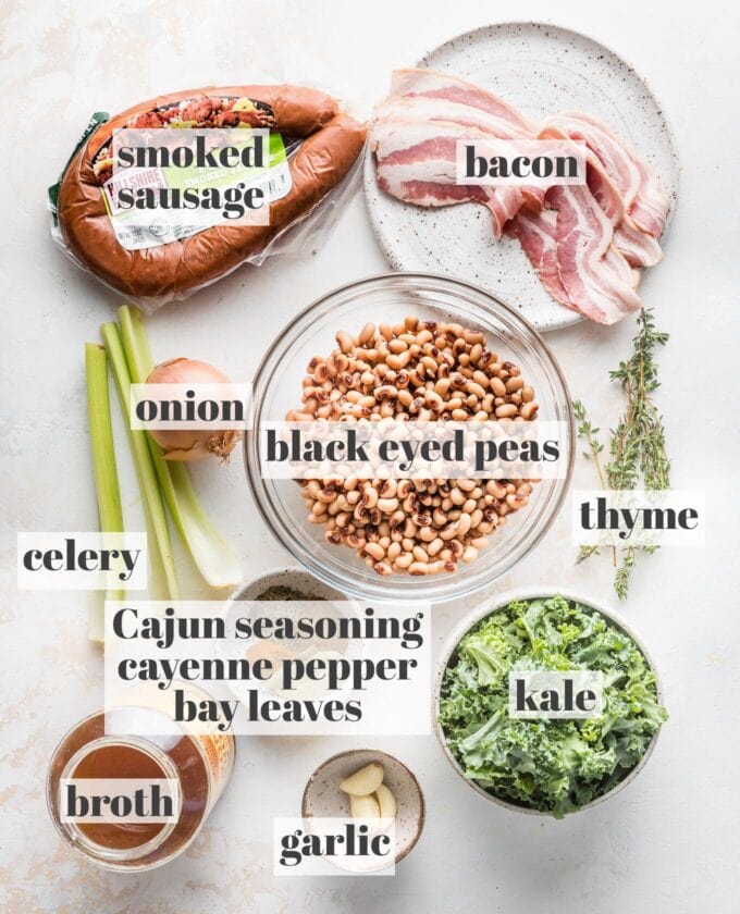 Labeled overhead photo of turkey smoked sausage, bacon slices, black eye peas, chopped kale, onion, garlic, broth, fresh thyme, and dried spices, laid out on a counter and ready to cook.