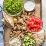Sheet pan filled with ingredients for chicken gyros: seasoned sliced chicken thighs, pita bread, shredded lettuce, chopped tomatoes, Feta, Tzatziki sauce, and cucumbers.
