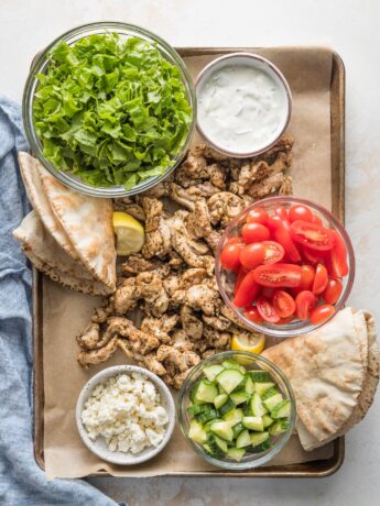 Sheet pan filled with ingredients for chicken gyros: seasoned sliced chicken thighs, pita bread, shredded lettuce, chopped tomatoes, Feta, Tzatziki sauce, and cucumbers.