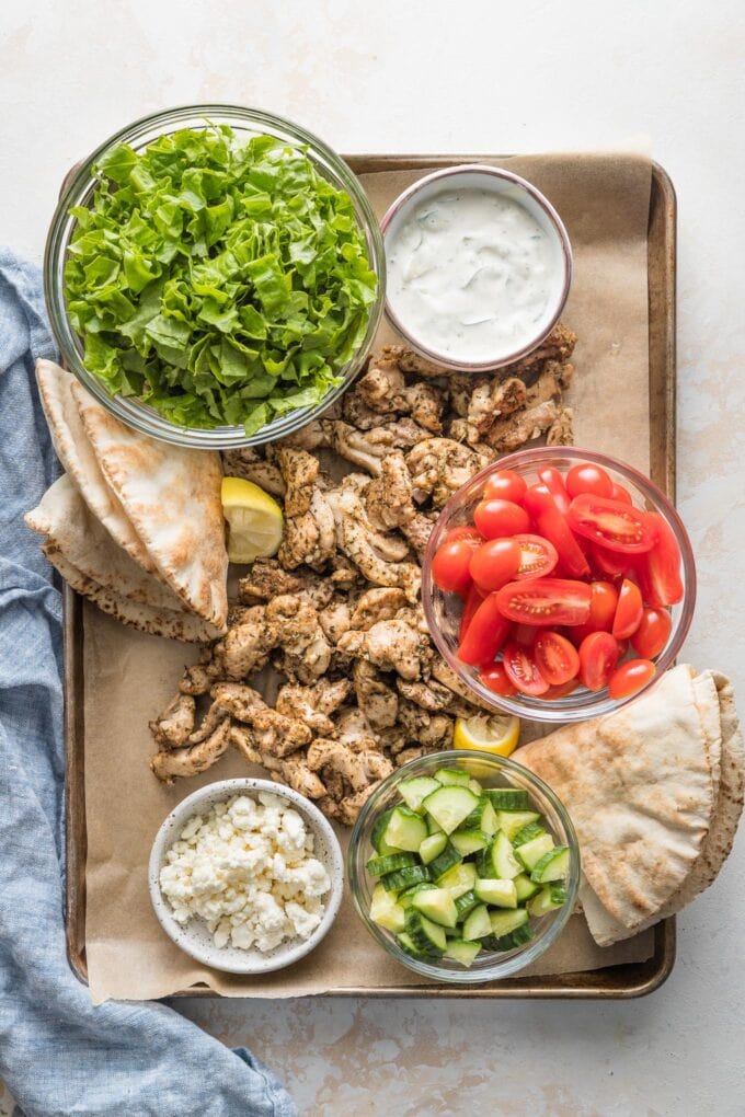 Sheet pan filled with ingredients for chicken gyros: seasoned sliced chicken thighs, pita bread, shredded lettuce, chopped tomatoes, Feta, Tzatziki sauce, and cucumbers.