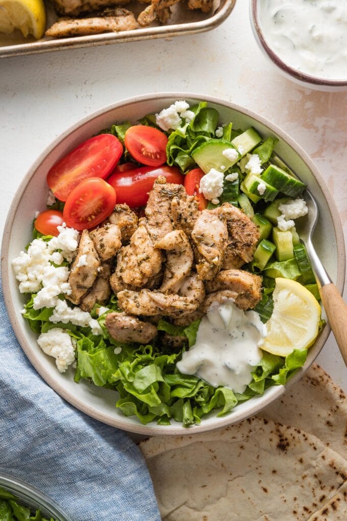 Bowl with a generous portion of sheet pan chicken gyros served with lettuce, tomato, cucumber, a lemon wedge, and Feta, with warmed pita on the side.