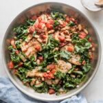 Skillet full of a Tuscan-inspired chicken dinner with kale, white beans, and sun-dried tomatoes, with salt and pepper in the background for added seasoning.