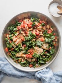 Skillet full of a Tuscan-inspired chicken dinner with kale, white beans, and sun-dried tomatoes, with salt and pepper in the background for added seasoning.