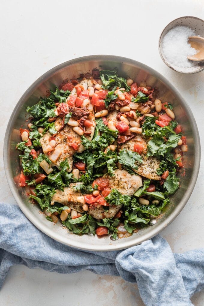 Skillet full of a Tuscan-inspired chicken dinner with kale, white beans, and sun-dried tomatoes, with salt and pepper in the background for added seasoning.