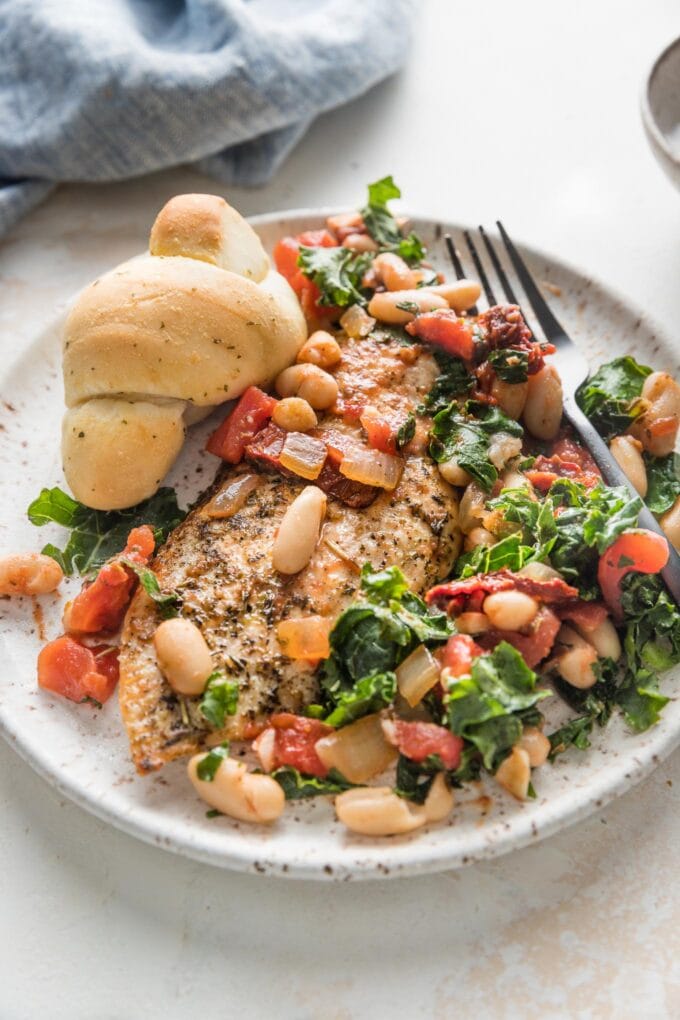 Small plate with a portion of chicken with white beans, kale, and sun-dried tomatoes, served with a garlic knot.