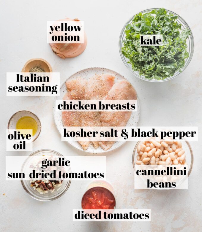 Labeled overhead photo of thin-sliced chicken breasts seasoned with salt and pepper, cannellini beans, diced tomatoes, minced garlic, sun-dried tomatoes, olive oil, Italian seasoning, yellow onion, and kale, all in prep bowls and ready to cook.