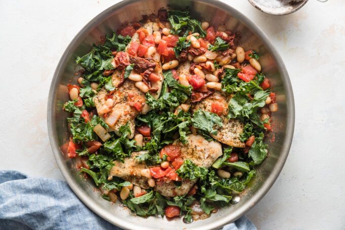 Skillet with chicken breasts, white beans, kale, and sun-dried tomatoes all in a light sauce.