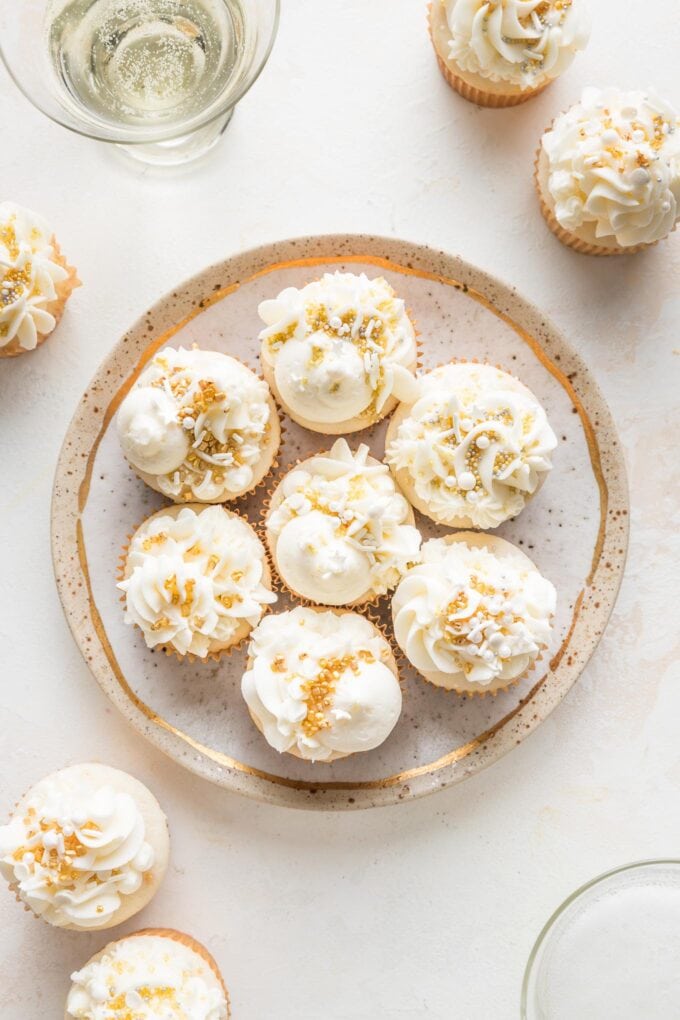 Countertop scattered with champagne cupcakes with buttercream, gold and white sprinkles, and couple glasses full of champagne.