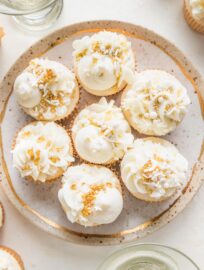 Small gold-rimmed plate filled with elaborately decorated champagne cupcakes, with white champagne buttercream and a mix of gold and white sprinkles.
