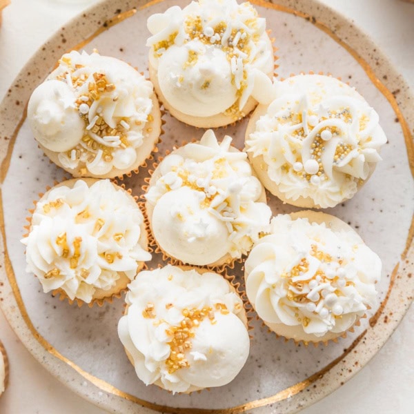 Small gold-rimmed plate filled with elaborately decorated champagne cupcakes, with white champagne buttercream and a mix of gold and white sprinkles.