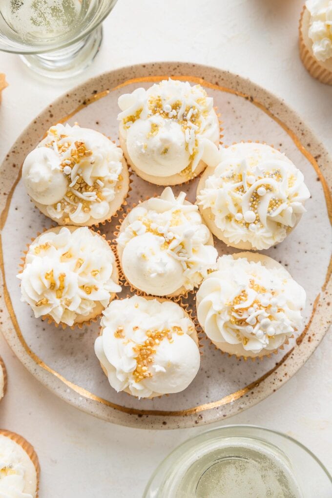 Small gold-rimmed plate filled with elaborately decorated champagne cupcakes, with white champagne buttercream and a mix of gold and white sprinkles.