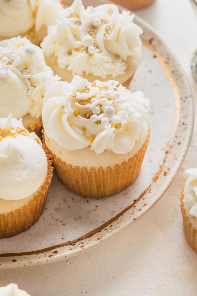 Close up angled view of a champagne cupcake decorated with champagne buttercream piped on in different designs and topped with gold and white assorted sprinkles.