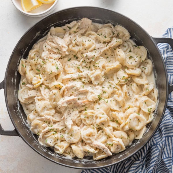 Large cast iron skillet full of creamy chicken tortellini Alfredo, with a blue kitchen towel and small bowls of lemon wedges and dried parsley scattered on the counter around it.