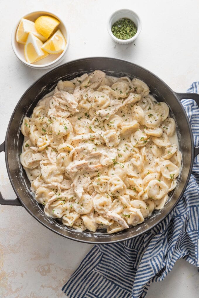 Large cast iron skillet full of creamy chicken tortellini Alfredo, with a blue kitchen towel and small bowls of lemon wedges and dried parsley scattered on the counter around it.