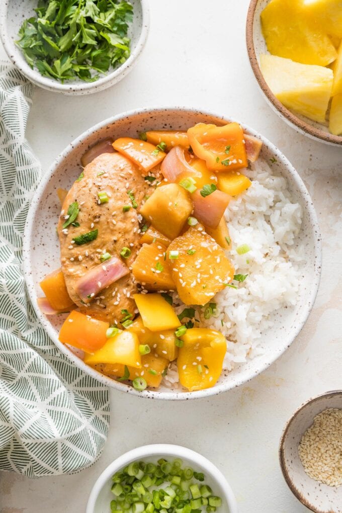 Bowl of Hawaiian-inspired pineapple chicken with peppers and onions served with white rice and garnished with sesame seeds, with extra pineapple chunks and garnishes in the background.
