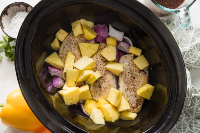 Seared chicken breasts in the bowl of a Crockpot with pineapple chunks and red onion.