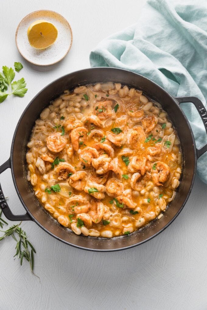 Large cast iron skillet full of a shrimp and bean stew garnished with fresh parsley.