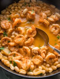 Close up of a serving spoon lifting a portion of rich, brothy shrimp stew out of a deep cast iron pan.