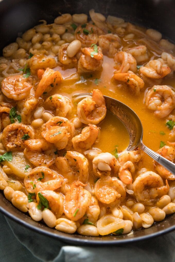 Close up of a serving spoon lifting a portion of rich, brothy shrimp stew out of a deep cast iron pan.