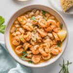 Overhead view of a shallow bowl filled with a shrimp and Cannellini bean stew, cooked in a rich, lemony rosemary broth, with garlic bread on the side.