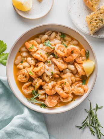 Overhead view of a shallow bowl filled with a shrimp and Cannellini bean stew, cooked in a rich, lemony rosemary broth, with garlic bread on the side.