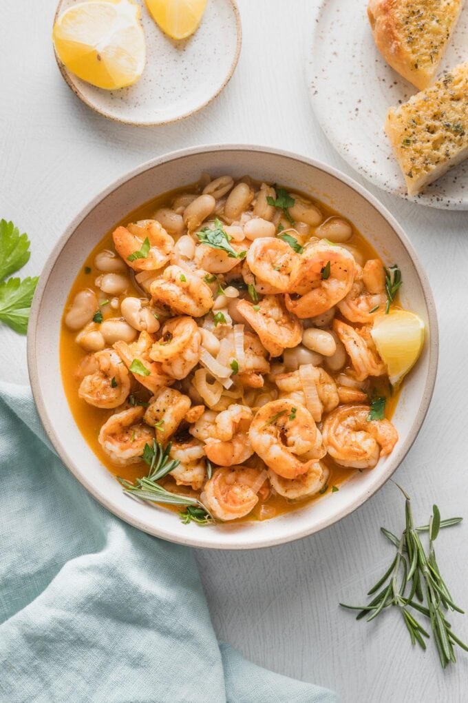 Overhead view of a shallow bowl filled with a shrimp and Cannellini bean stew, cooked in a rich, lemony rosemary broth, with garlic bread on the side.