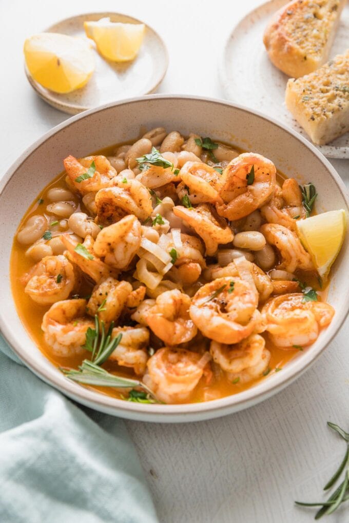 Angled view of lemony shrimp and bean stew in a shallow serving bowl with garlic bread and extra lemon wedges on the side.