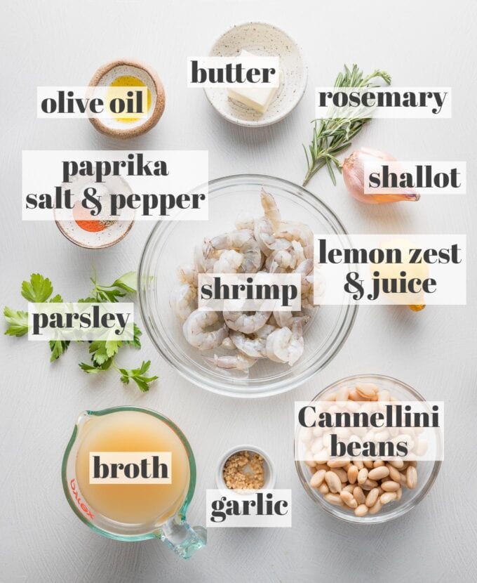 Labeled overhead photo of raw shrimp, Cannellini beans, broth, minced garlic, a whole lemon, a whole shallot, sprigs of fresh rosemary and parsley, butter, olive oil, paprika, salt, and pepper, all measured into prep bowls and ready to cook.