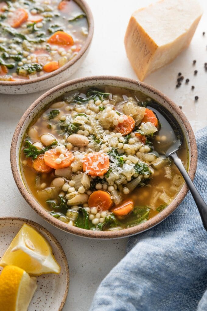 Angled view of a soup bowl filled with a brothy mixture of white beans, pearl couscous, Parmesan, and veggies.