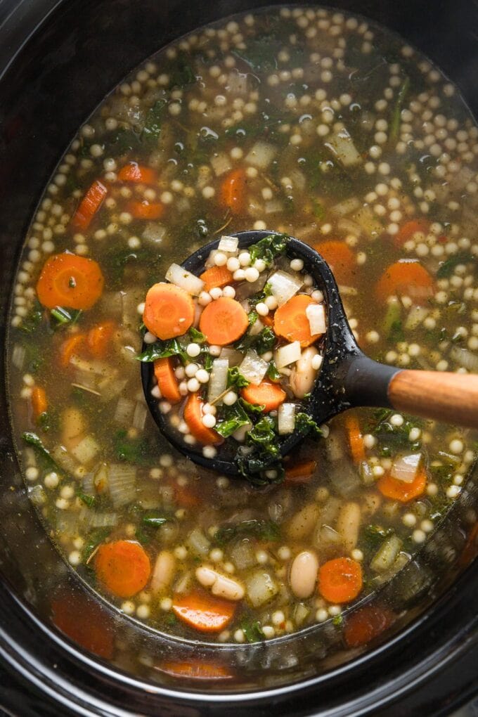 Close up view of a ladle lifting a portion of white bean Parmesan soup out of a large black Crockpot.