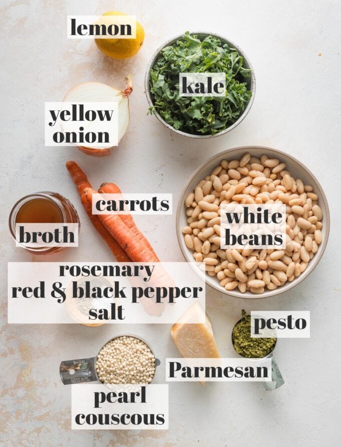 Labeled overhead photo of white Cannellini beans, carrots, onion, kale, pesto, Parmesan, a lemon, chicken broth, pearl couscous, dried rosemary, red and black pepper, and salt all measured and ready to cook.
