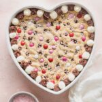 Heart shaped chocolate chip cookie cake decorated with frosting, pink and white M&Ms, and pink sprinkles.