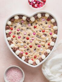 Heart shaped chocolate chip cookie cake decorated with frosting, pink and white M&Ms, and pink sprinkles.