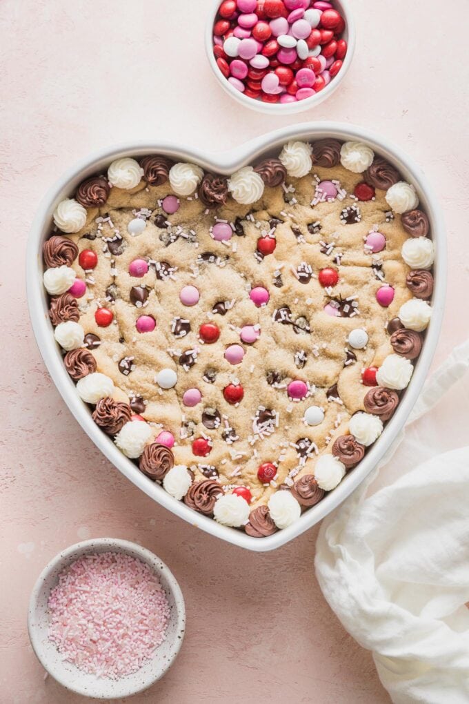 Heart shaped chocolate chip cookie cake decorated with frosting, pink and white M&Ms, and pink sprinkles.