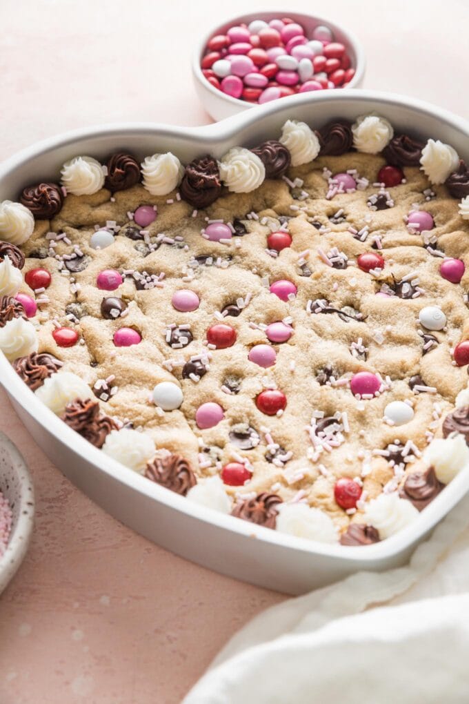 Angled view of a heart-shaped chocolate chip cookie cake being decorated with chocolate and vanilla frosting.