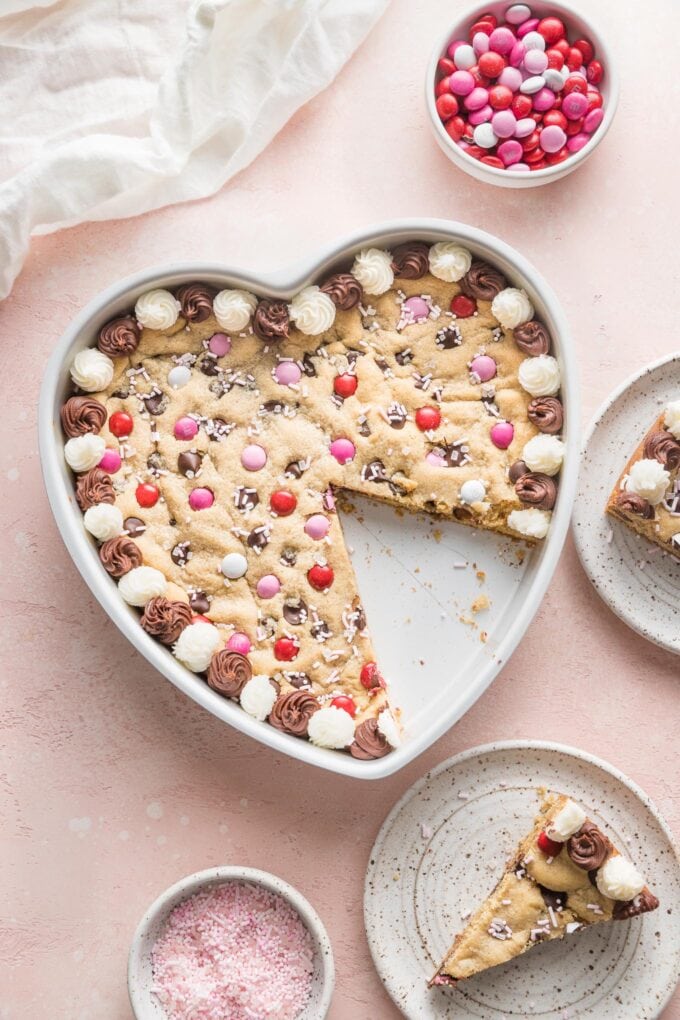 Overhead view of a heart-shaped cookie cake with several clean wedges cut and lifted out to serve on small dessert plates.