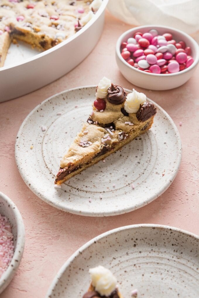 Angled view of a slice of cookie cake served on a small plate with extra M&Ms in the background.