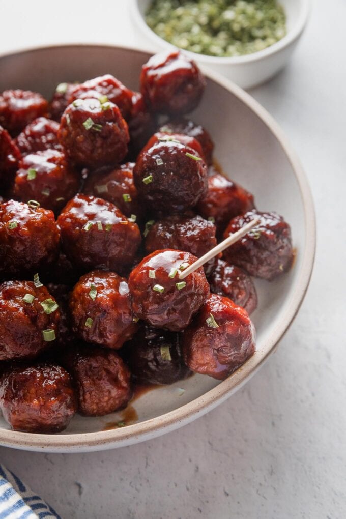 Close up of a slow cooker BBQ meatball with a toothpick in it, ready to be picked up and eaten out of a ceramic bowl.